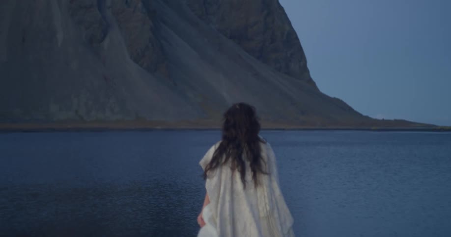 A mysterious woman with long dark hair in a flowing white gown slowly walks towards a dark lake in a dramatic mountain landscape at dusk