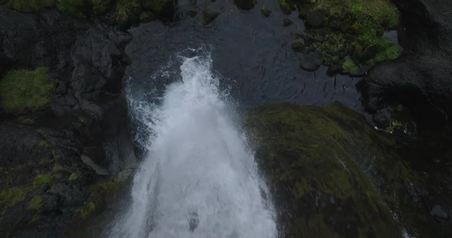 A slow-motion aerial view glides over the edge of a beautiful waterfall in a lush, moss-covered canyon