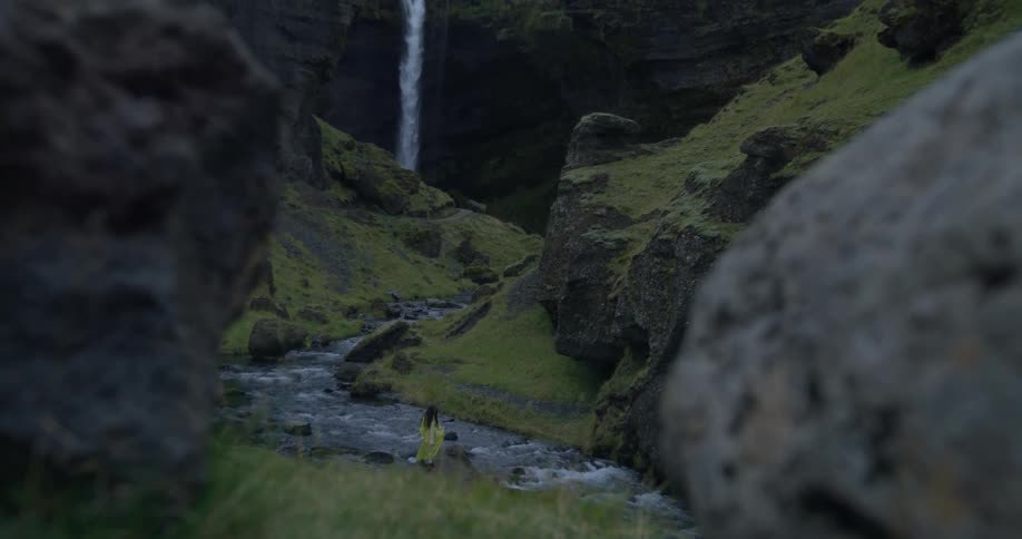 A person in a bright yellow raincoat stands on a rock next to a flowing river, admiring a majestic waterfall in a lush green canyon