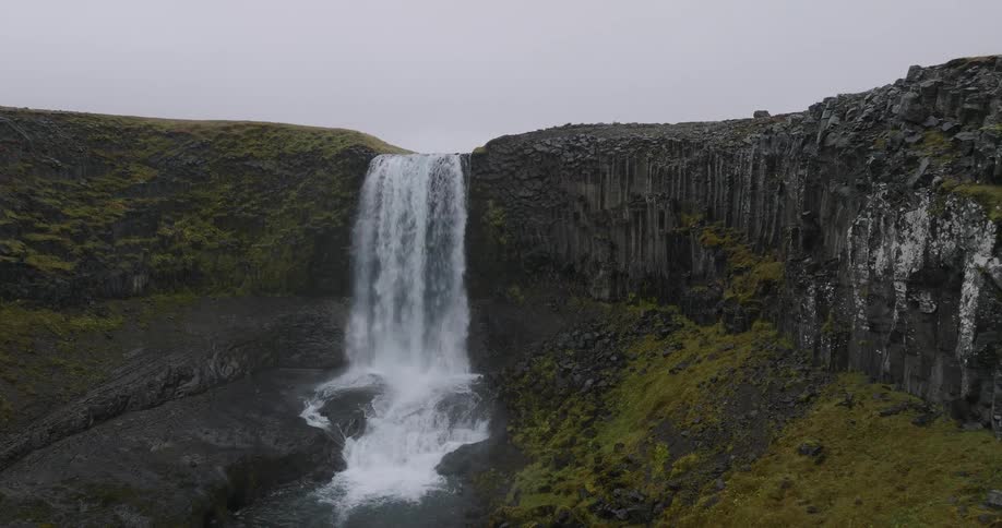 An aerial view slowly approaches a majestic waterfall cascading over dramatic basalt rock columns in a rugged Icelandic landscape