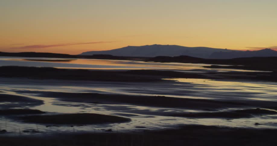 A stunning sunset casts a warm glow over a tranquil tidal estuary with snow-capped mountains in the distance