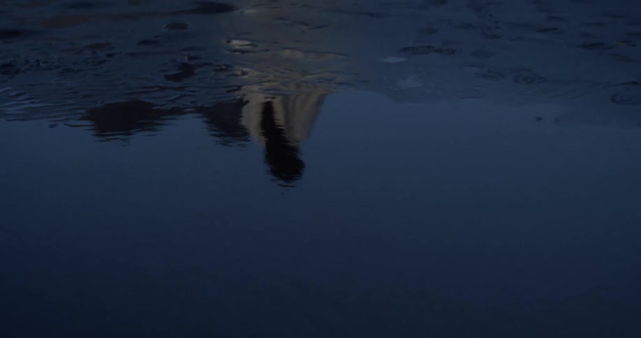 The distorted reflection of a robed figure disappears from a puddle on a sandy beach as they walk away at dusk