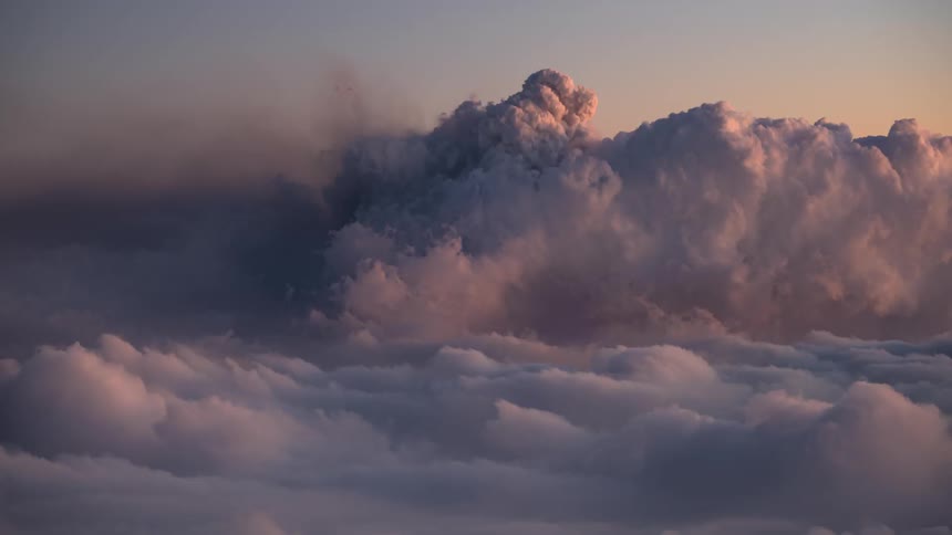 A dramatic day-to-night timelapse captures a volcano erupting above a sea of clouds, with the ash plume illuminated by the setting sun and the glowing lava at night