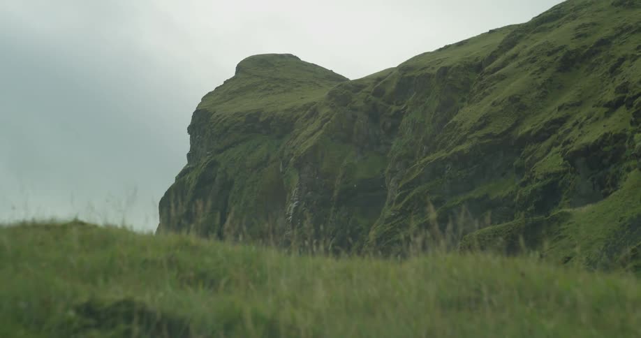 A close-up of lush green grass on a hillside slowly reveals a majestic rocky cliff in the background