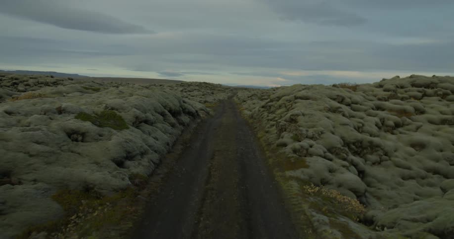 An aerial shot slowly pulls back along a dirt road that cuts through a vast, otherworldly landscape of moss-covered volcanic rocks in Iceland