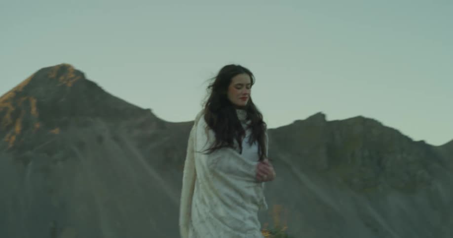 A pregnant woman in a white dress and shawl walks along a black sand beach with majestic mountains in the background