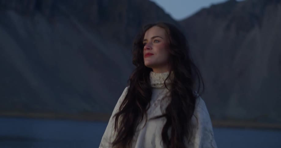A serene woman in a flowing white dress and lace shawl walks in slow motion along a tranquil shore with mountains in the background at dusk