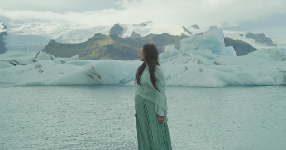 A pregnant woman in a flowing green dress walks to the edge of a glacial lagoon, peacefully holding her belly while surrounded by icebergs and snowy mountains