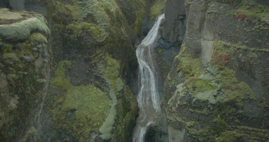 A stunning high-angle shot follows a winding waterfall as it cascades through a lush, moss-covered canyon