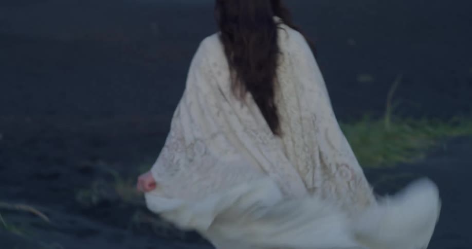 A woman with long dark hair in a flowing white lace dress walks in slow motion on a dark sand beach at dusk