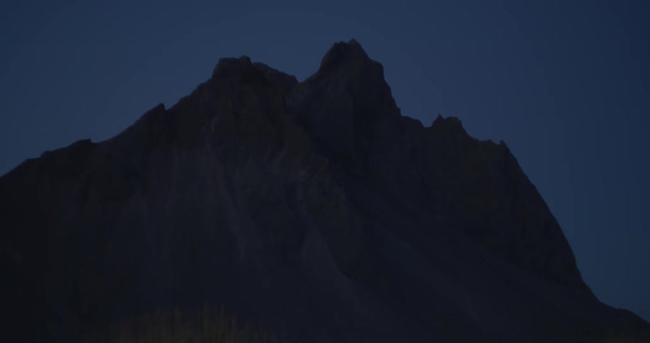 The jagged peaks of a mountain are silhouetted against a deep blue sky during twilight