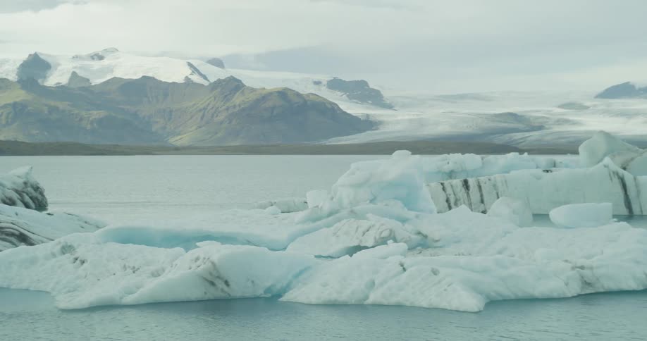 A slow pan reveals majestic icebergs floating in a glacial lagoon with snow-capped mountains and a massive glacier in the background