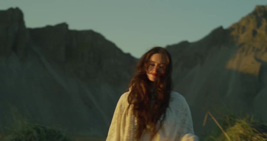 A beautiful woman in a flowing white lace dress walks towards the camera in slow motion, with majestic mountains in the background during a golden sunset
