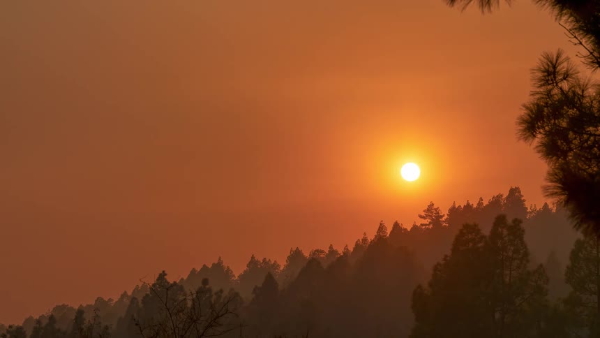 The brilliant sun hangs in a hazy orange sky over a silhouetted pine forest on a mountainside