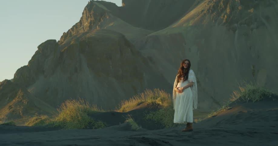 A contemplative pregnant woman in a white dress stands on a black sand beach in a dramatic landscape during the golden hour