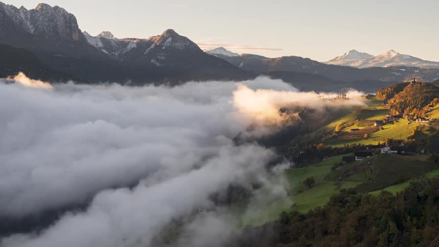 A stunning timelapse captures the sunrise over a foggy mountain valley as the clouds slowly dissipate to reveal the green hills and a small village below