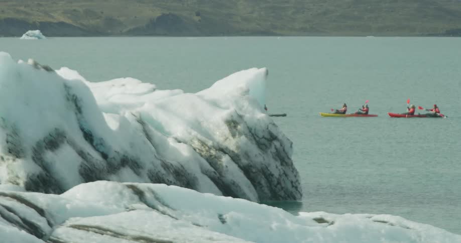 A group of kayakers paddles through a calm glacial lagoon surrounded by massive icebergs