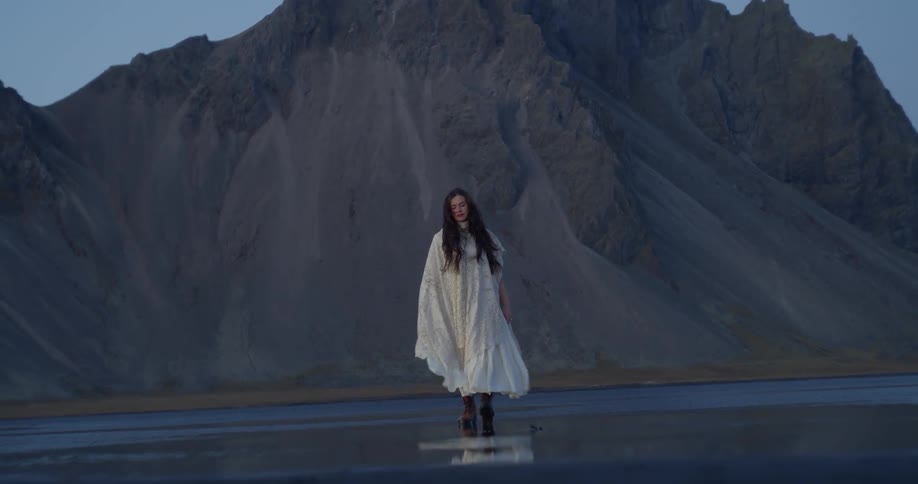 An ethereal woman in a flowing white dress and cape walks in slow motion across a reflective, dark beach in front of a dramatic Icelandic mountain