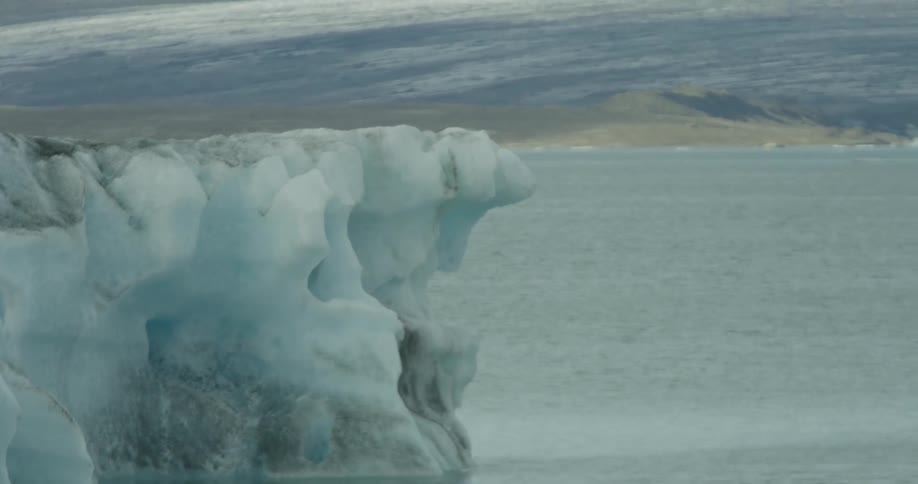 A slow pan across a large, detailed blue iceberg floating in a calm glacial lagoon reveals a vast glacier in the background