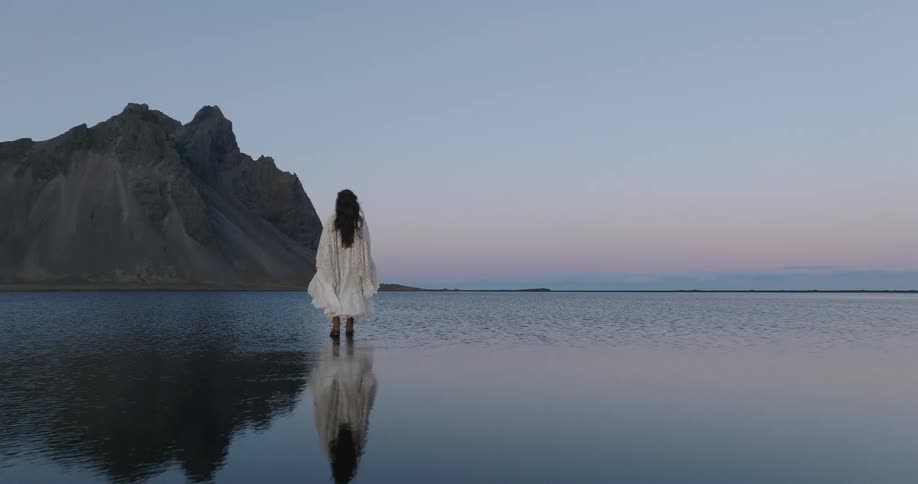 A serene woman in a flowing white dress stands in the tranquil waters of a lagoon before a majestic mountain at twilight