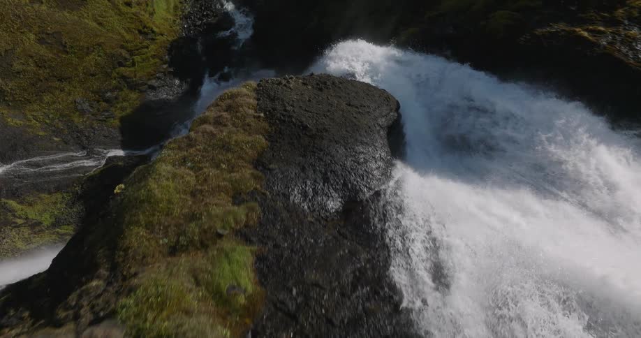 Slow aerial push-in over the edge of a powerful waterfall cascading down a mossy, rocky cliff