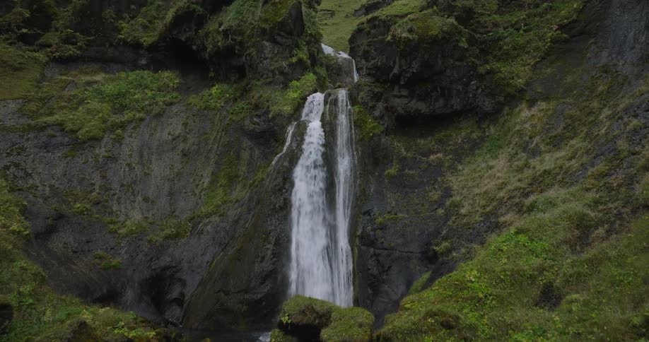A drone slowly pushes in on a beautiful cascading waterfall in a lush, mossy green canyon