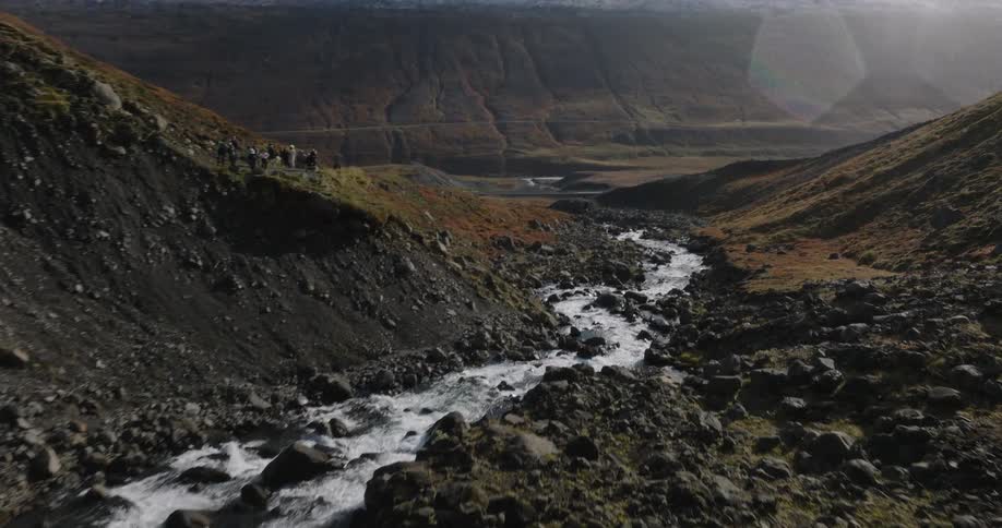 A slow-motion drone shot tilts up from a rushing rocky river to reveal a vast, mountainous landscape with snow-capped peaks in the distance