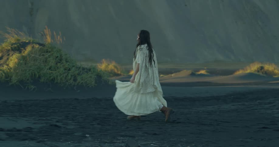 A woman in a flowing white dress walks in slow motion across a desolate black sand beach against a backdrop of mountains