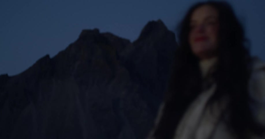 A woman with windblown hair smiles peacefully at dusk, with a rack focus from a jagged mountain peak