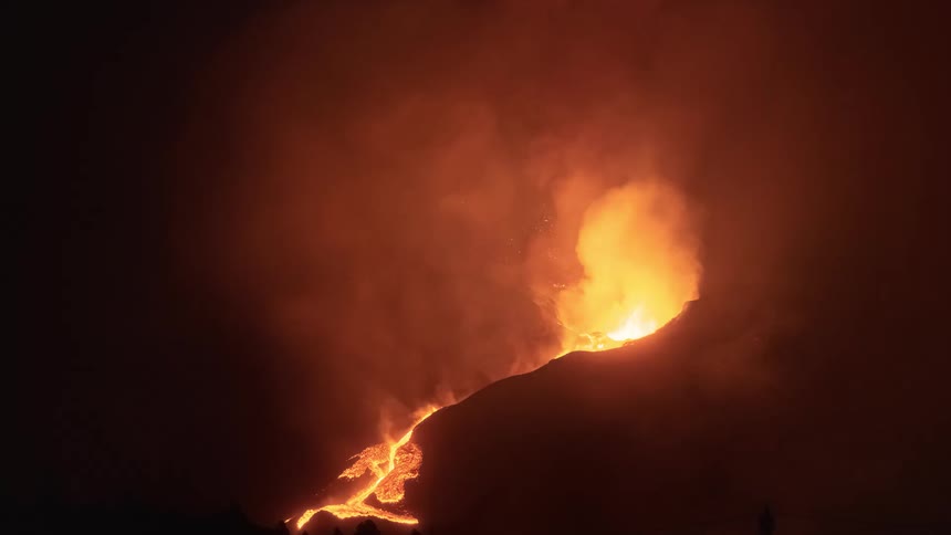 A volcano erupts with fiery explosions at night, spewing molten lava down its slopes under a thick cloud of smoke and ash