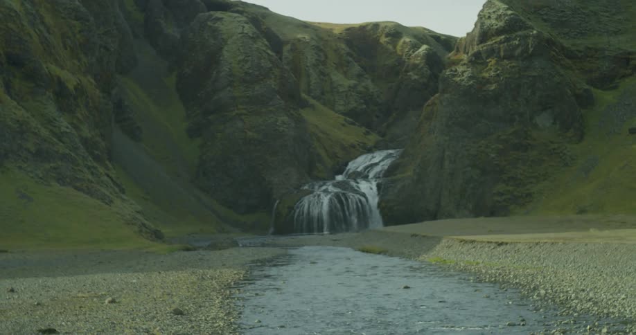 A slow reveal of a picturesque waterfall cascading down mossy green cliffs into a river in a remote Icelandic valley