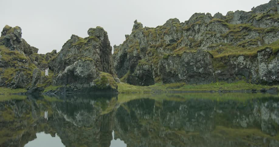 A woman in a white dress stands on a mossy, volcanic cliff overlooking a still lake that perfectly reflects the rugged landscape