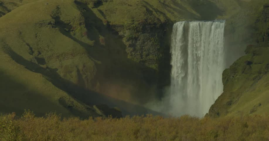 A pan reveals the magnificent Skogafoss waterfall cascading down a verdant cliffside in Iceland, with a faint rainbow in the mist at its base