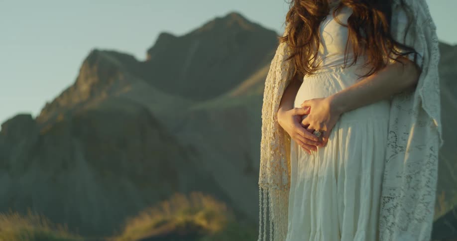 A close-up shot of a pregnant woman in a white dress holding her belly in front of a mountain range during a golden hour sunset