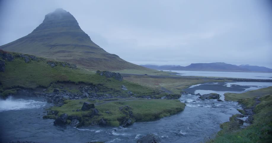 A slow pan reveals the stunning Kirkjufellsfoss waterfall and the iconic Kirkjufell mountain in Iceland under a cloudy sky