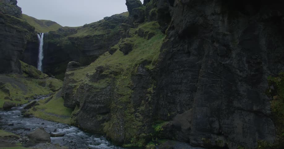 A dramatic pan away from a moss-covered cliff reveals a powerful waterfall flowing into a river in a majestic Iceland canyon