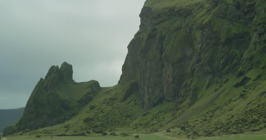 A slow panning reveal of a dramatic, moss-covered cliff rising above a green field in the Icelandic wilderness