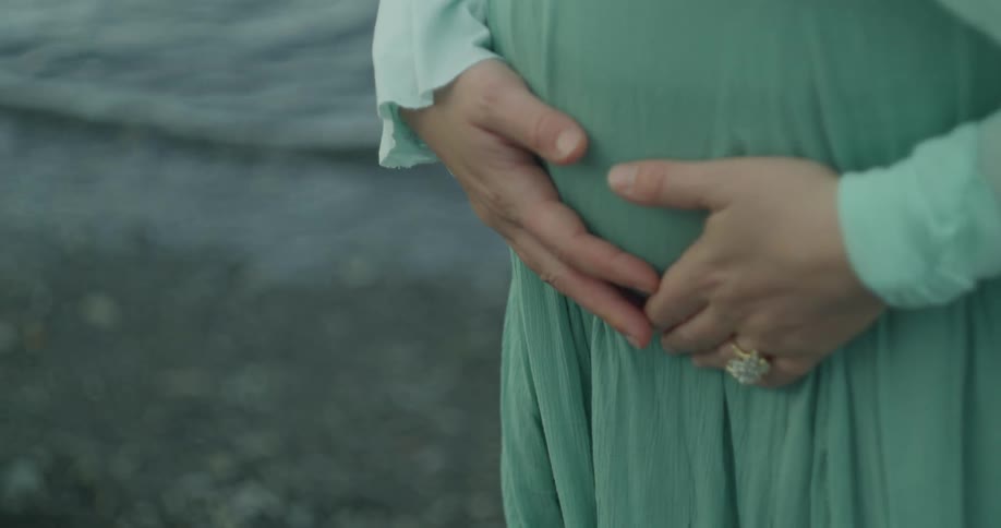 A close-up shot shows a pregnant woman in a flowing green dress making a heart shape with her hands over her baby bump near the water