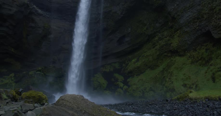 A river flows over mossy rocks before the camera pans up to reveal a majestic waterfall crashing down a dramatic cliffside