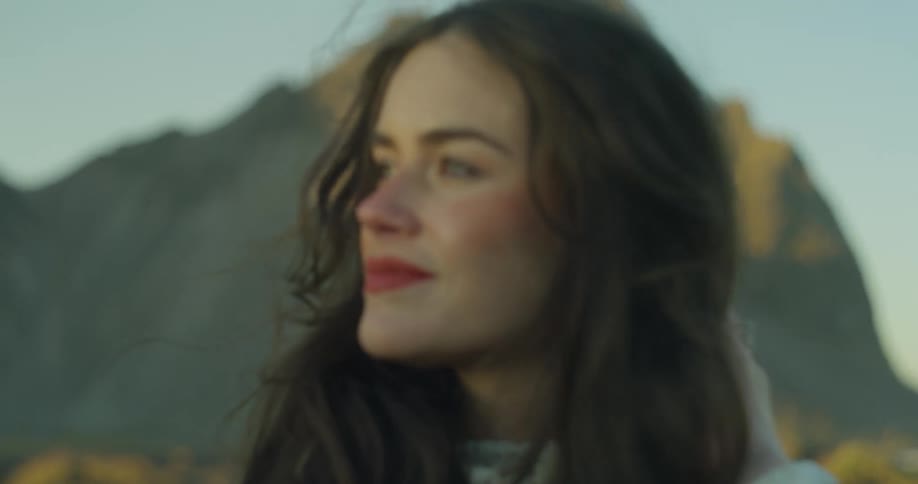 A cinematic slow-motion close-up of a thoughtful young woman with her hair blowing in the wind, set against a majestic mountain range during the golden hour