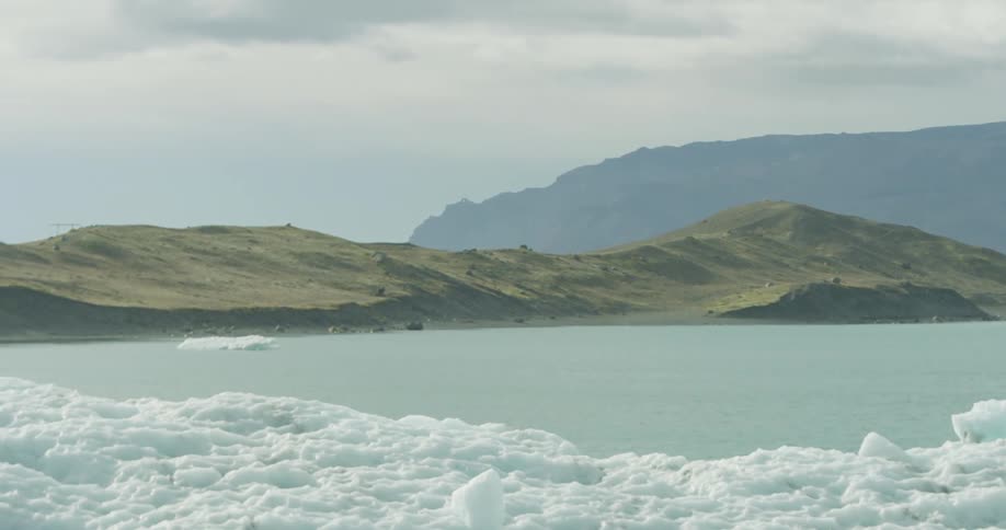A white bird soars in slow motion over an icy glacier lagoon with snow-capped mountains in the background