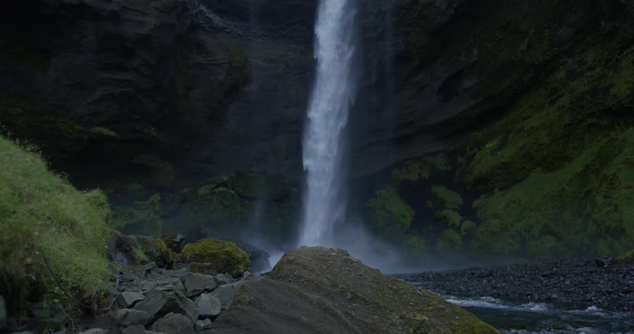 Tilting up from the rocky riverbed to reveal a powerful waterfall crashing down from a mossy cliff in Iceland