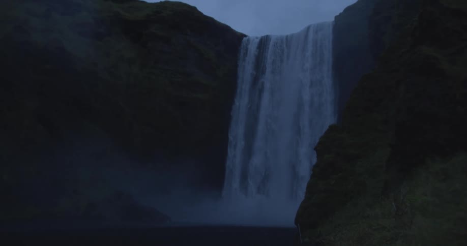 A powerful, misty waterfall plunges over a dark, moss-covered cliff in a moody, low-light shot