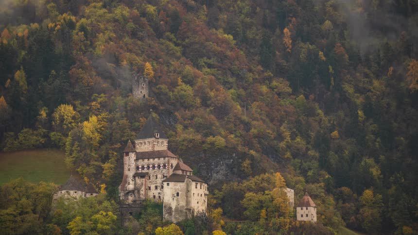 Timelapse of misty clouds rolling over a historic castle nestled in a colorful autumn forest on a mountainside
