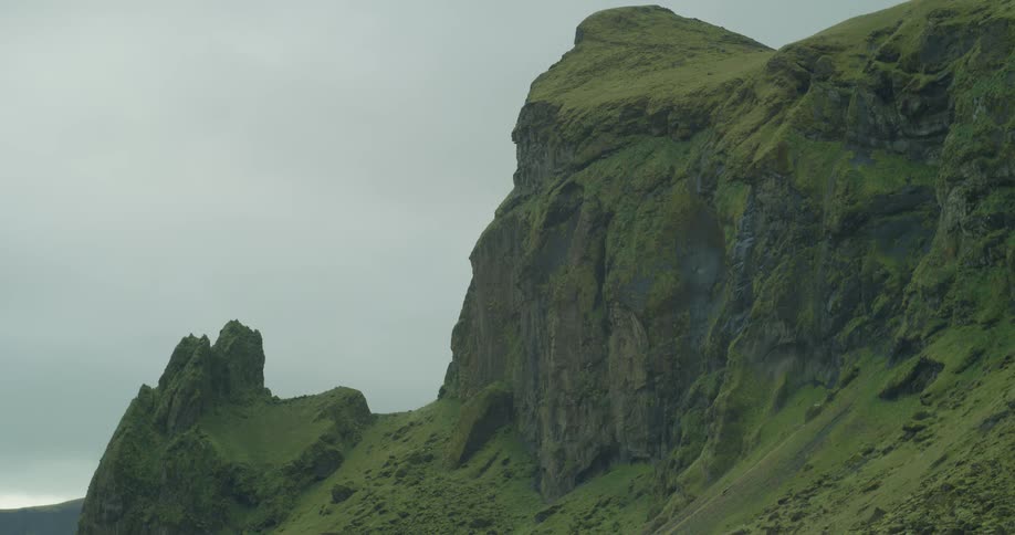 A towering, moss-covered cliff face stands against a vast, overcast sky in a rugged, green landscape