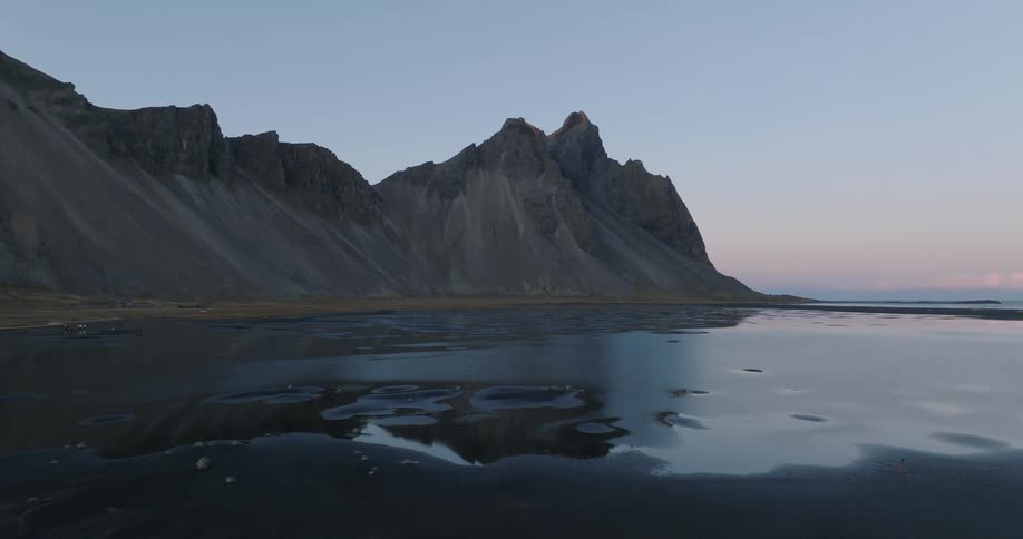 The majestic Vestrahorn mountain range is perfectly reflected in the calm waters of a black sand beach in Iceland during a serene twilight pan shot