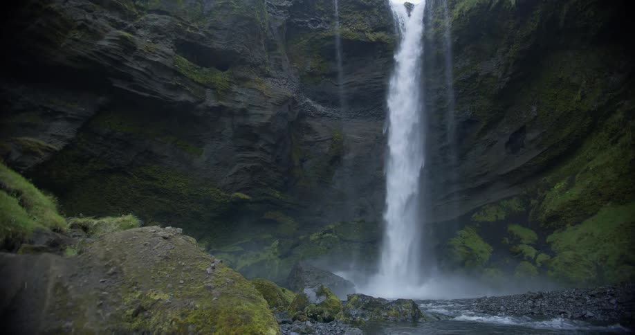 A powerful waterfall crashes into a rocky stream at the base of a dark, mossy canyon