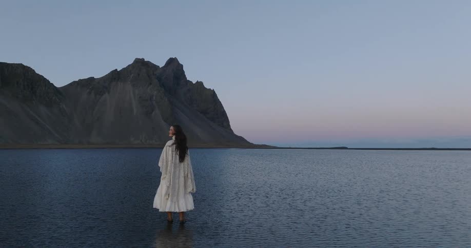 A solitary woman in a flowing white dress and coat contemplates the serene landscape of a mountain and lake at twilight