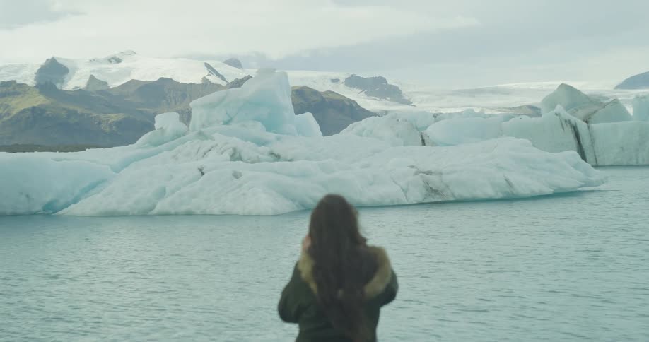 A woman with long brown hair stands still, observing the awe-inspiring sight of large icebergs in a glacial lagoon