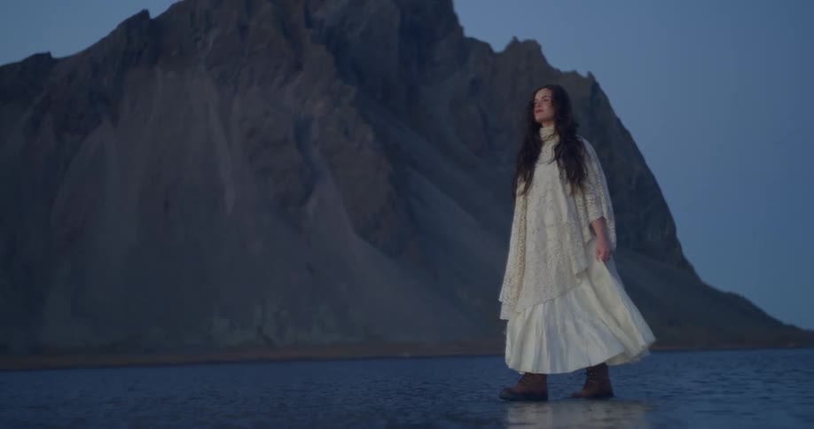 A woman in a flowing white vintage dress walks through the shallow water before the majestic Vestrahorn mountains in Iceland at dusk
